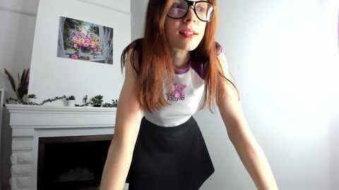 Media: Video of a Caucasian woman with long red hair, wearing black-framed glasses, a white tank top with a pink graphic, and a black skirt, standing indoors near a white fireplace with a floral picture.