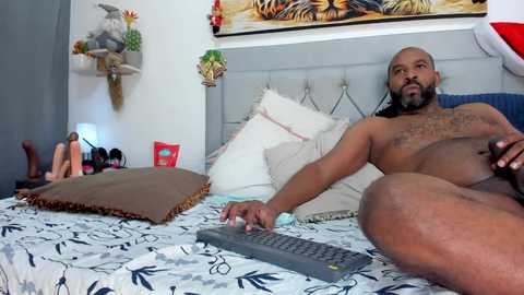 Media: A video of a nude Black man with a muscular build and medium skin tone, lounging on a bed with white and black patterned sheets. He holds a remote control. The room features colorful decor and a TV on the wall.