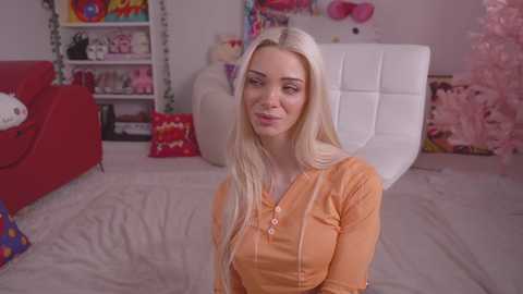 Media: Video of a young, blonde, fair-skinned woman with a slim figure and medium-sized breasts, wearing an orange top, sitting on a bed in a colorful, themed room.