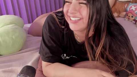 Media: Video of a smiling woman with long dark hair, wearing a black t-shirt, lying on a bed with a green pillow, purple wall in the background.