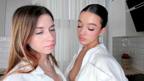 Media: Video of two young women with light skin, one with long, wavy, brown hair and the other with dark hair in a bun, wearing white robes, in a modern, white-tiled kitchen.