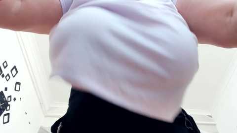 Media: A close-up video of a person wearing a tight, white, short-sleeved shirt and black pants, with a blurred background featuring geometric patterns and white walls.