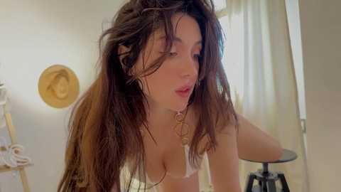 Media: Video of a young, slender, light-skinned woman with long, wavy brown hair, wearing a white bra, leaning forward. Background shows a room with a ladder, a hat, and a chair.