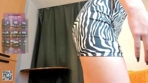 Video of a person wearing a tight, zebra-print tank top, standing in a room with a green curtain, wooden shelf, and a chart displaying weight loss progress.