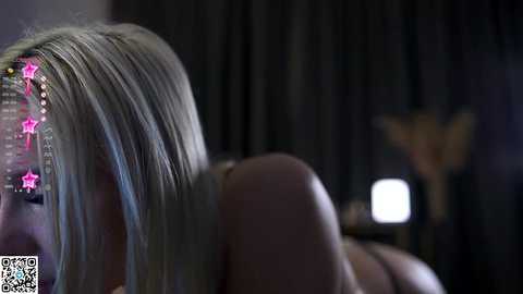 Media: A dimly lit video of a blonde woman with long, straight hair, wearing a starry headband, lying on her side. The background is dark with blurred elements.