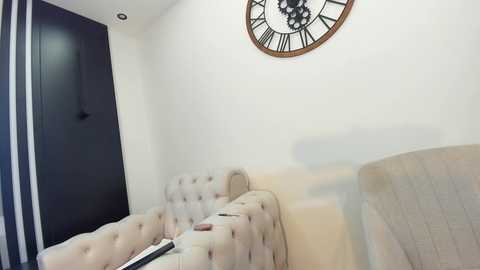 Media: A video of a minimalist, modern bedroom with a beige tufted headboard, white walls, and a large, round clock above a black door.