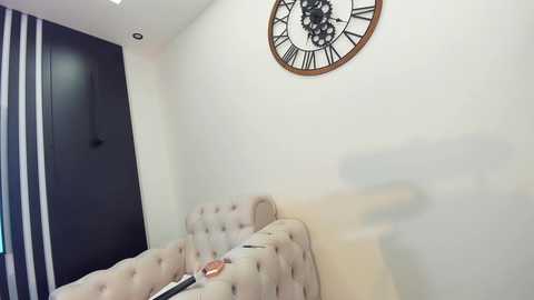 Media: A video of a modern, minimalist bedroom with a beige tufted headboard, a large wall clock, and vertical striped curtains.