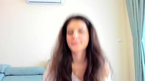 Media: A blurry video of a woman with long brown hair, wearing a light-colored top, standing in a room with a blue couch, white wall, and teal curtains.