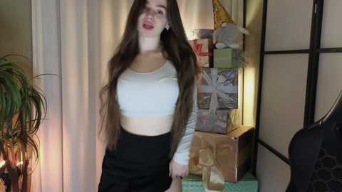 Media: A video of a young woman with long brown hair, wearing a white crop top and black high-waisted pants, standing in a cozy room with festive decorations and a plant.