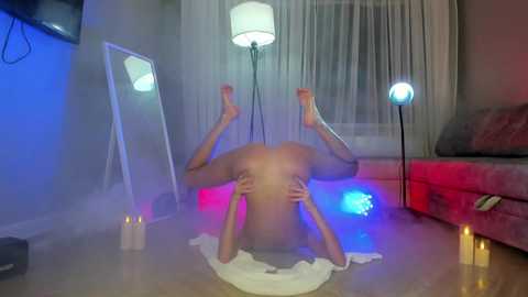 Media: Video of a nude, slender woman in a yoga pose on a carpet, surrounded by candles and colorful lights, in a dimly lit living room.