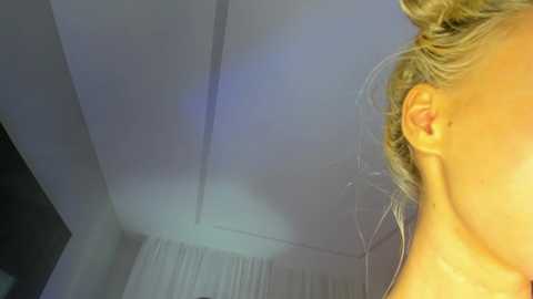 Media: A video captures a blonde woman's partially visible head and upper back, with her hair tied back. The background features a white ceiling and a hint of a curtain.