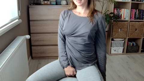 Media: Video of a light-skinned woman with brown hair, sitting on a chair in a room with wooden furniture and bookshelves. She wears a gray long-sleeve shirt and light blue pants, smiling.