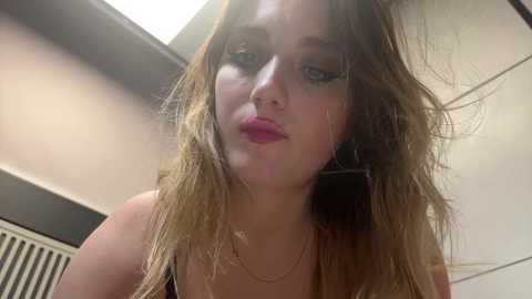 Media: Video of a young woman with long, messy blonde hair, wearing a black top, standing indoors with a ceiling light above. She has fair skin, blue eyes, and a slight smile.