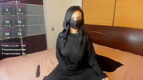 Media: Video of a Black woman with long black hair, wearing a black face mask, black long-sleeve top, and black pants, sitting on a bed with a wooden headboard, in a dimly lit bedroom.