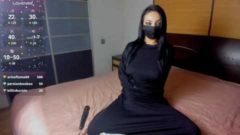 Media: A video of a woman with long black hair, wearing a black face mask, black dress, and black gloves, sitting on a bed in a dimly lit bedroom.