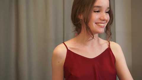 Media: Video of a young woman with fair skin and shoulder-length brown hair, smiling. She wears a sheer, red camisole with thin straps. Background shows blurred beige curtains.