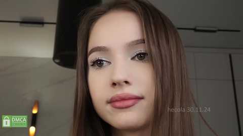 Media: Video of a young woman with long brown hair, light skin, and subtle makeup, standing in a modern kitchen with a white cabinet and dim lights.