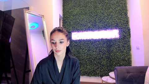 Media: Video of a young woman with fair skin, dark hair in a ponytail, wearing a black robe, standing in a modern salon with a green wall and a glowing neon sign.