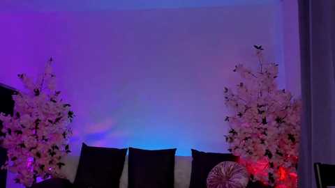 Media: Video of a stage with purple and blue lighting, featuring two tall floral arrangements with pink and white blooms on either side, black curtains, and a large circular fan at the center.