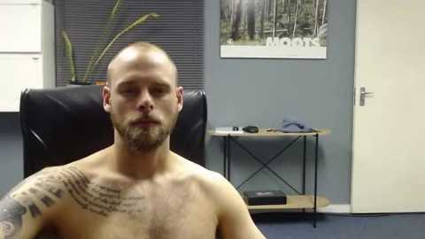 Media: Video of a shirtless, muscular, bald man with a beard and tattoos, seated in an office with a black chair, grey walls, and a \"moons\" print on the wall.