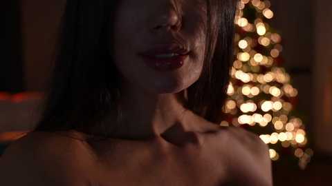 Media: Video of a woman with fair skin and long, dark hair, partially illuminated by warm, bokeh Christmas lights in the background.