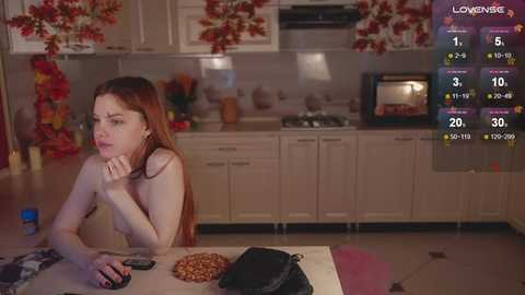 Media: Video of a nude, slender, red-haired woman seated at a kitchen table, eating cookies, surrounded by floral-patterned cabinets, and a stove in the background.