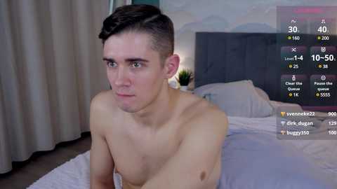 Media: Video of a young, shirtless, fair-skinned man with short brown hair, sitting on a bed in a modern bedroom. He has a serious expression.