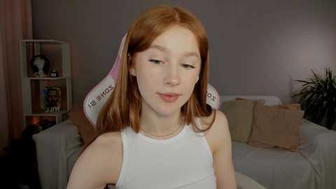 Video of a young, fair-skinned woman with red hair, wearing a white tank top, sitting in a dimly lit living room with a white couch, beige pillows, and a shelf with books and decor.