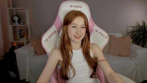 Media: Video of a young Caucasian woman with long red hair, wearing a white tank top, seated in a pink and white gaming chair with \"Gaming Chair\" text. Background shows a beige couch, white shelves, and a green plant.