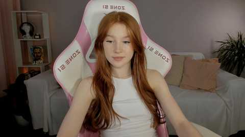 Video of a fair-skinned, red-haired woman in a white tank top, sitting in a pink gaming chair, with a beige sofa and shelves in the background.