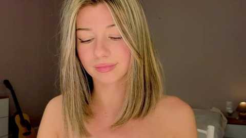 Media: Video of a young Caucasian woman with straight, shoulder-length blonde hair, fair skin, and closed eyes, wearing no shirt, in a dimly lit bedroom with a gray bed, a guitar, and a nightstand.