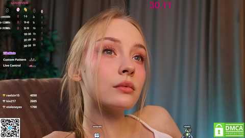 Media: Video of a young Caucasian woman with fair skin, blonde hair, and light makeup, wearing a white tank top, seated on a brown couch. Background includes a streaming platform with a timestamp and chat box.