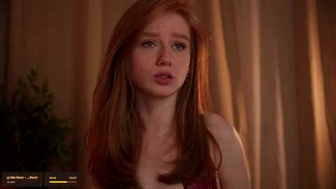 Media: Video of a young woman with long, straight, reddish-brown hair, fair skin, and blue eyes, wearing a sleeveless red top, against a warm, textured curtain background.