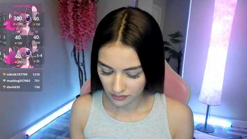 Media: Video of a young woman with straight, dark brown hair in a light gray tank top, seated in a pink chair. Background features a pink floral arrangement, a large white lamp, and a digital screen displaying social media icons.