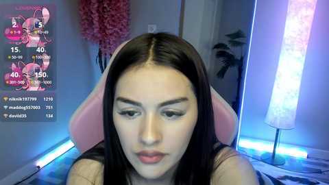 Media: A video of a young woman with long dark hair in a pink gaming chair, looking at a computer screen with a chat window. Background includes a pink tree and a tall light.