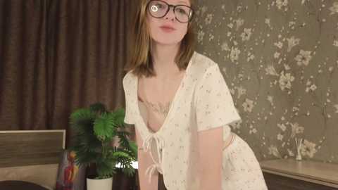 Media: Video of a young Caucasian woman with straight, light brown hair, wearing black-rimmed glasses, a white floral dress, and a potted green plant. Background includes brown curtains and patterned wallpaper.