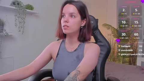 Media: A video of a young woman with shoulder-length dark hair, fair skin, and a gray tank top, sitting in a black gaming chair. The background features a potted plant and a digital screen displaying gaming stats.