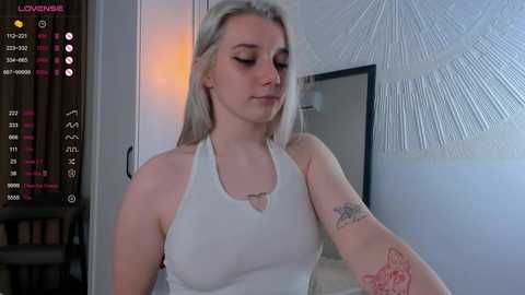 Media: Video of a young, fair-skinned woman with long, straight platinum blonde hair, wearing a white tank top, standing in a dimly lit room with a wall-mounted mirror and a starburst pattern.