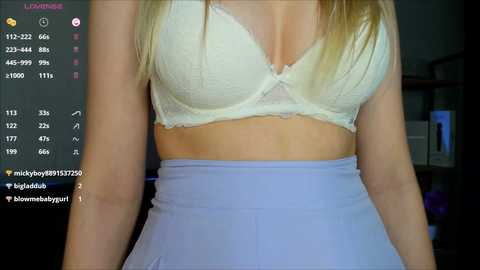 Media: A video of a fair-skinned woman with long blonde hair, wearing a white lace bra and a high-waisted, light blue skirt, indoors with a dark background, with social media icons and text in the top left corner.