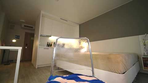 Media: Video of a minimalist, modern bedroom with a white bed, beige bedding, and a clear chair with a blue patterned cushion. The room features white cabinets and a kitchenette with bottles, a small table, and a round lamp.