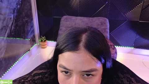 Media: Video of a woman with straight black hair, wearing a black lace top, seated at a table with a textured black wall and green LED lights in the background.