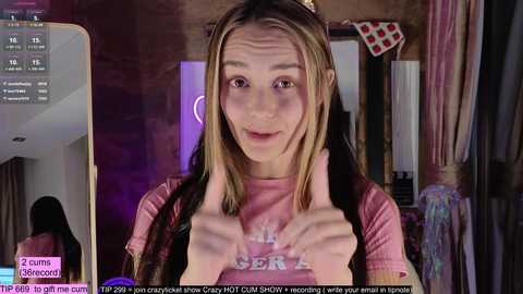 Media: A video of a young woman with long, straight blonde hair, wearing a pink T-shirt, making a heart with her hands in a cozy, dimly lit room.