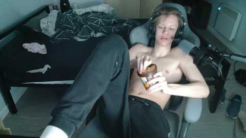 Media: A candid video of a shirtless young man with pale skin and light brown hair, wearing headphones, drinking from a can on a gaming chair in a cluttered room with a bed, clothes, and gaming equipment.