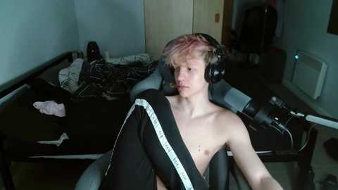 Media: Video of a shirtless, young, fair-skinned boy with pink hair, wearing headphones, sitting in a gaming chair in a cluttered, dimly lit room.