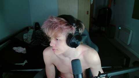 Media: A young, fair-skinned, shirtless man with messy pink hair, wearing large black headphones, sits in a dimly lit room with cluttered furniture, including a bed with disheveled bedding, and a gaming chair.