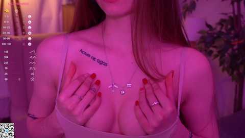 Media: Video of a light-skinned woman with long red hair, wearing a sheer, white top, covering her breasts with her hands. She has a tattoo on her chest. Background features a potted plant.