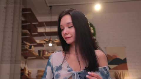 Media: Video of a young Asian woman with long black hair, wearing a tie-dye top, standing in a modern, warmly lit room with exposed brick walls, wooden shelves, and a colorful abstract painting.