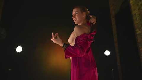 Media: Video of a fair-skinned woman with long brown hair, wearing a pink satin gown, adjusting her hair, in a dimly lit outdoor setting with two bright spotlights in the background.