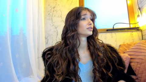 Media: Video of a young woman with long, wavy brown hair, wearing a black fur coat, standing in a dimly lit room with a beige wall and a window with a light curtain.