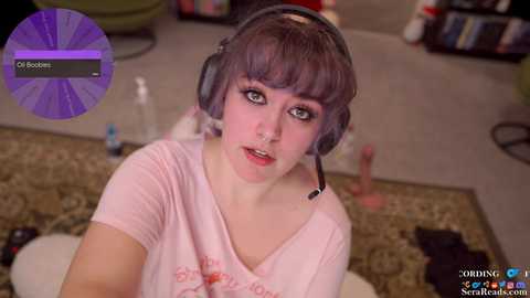 Video of a young woman with pale skin, short dark hair, and wearing a headset, sitting on a patterned rug in a cozy room with a bookshelf and scattered items.